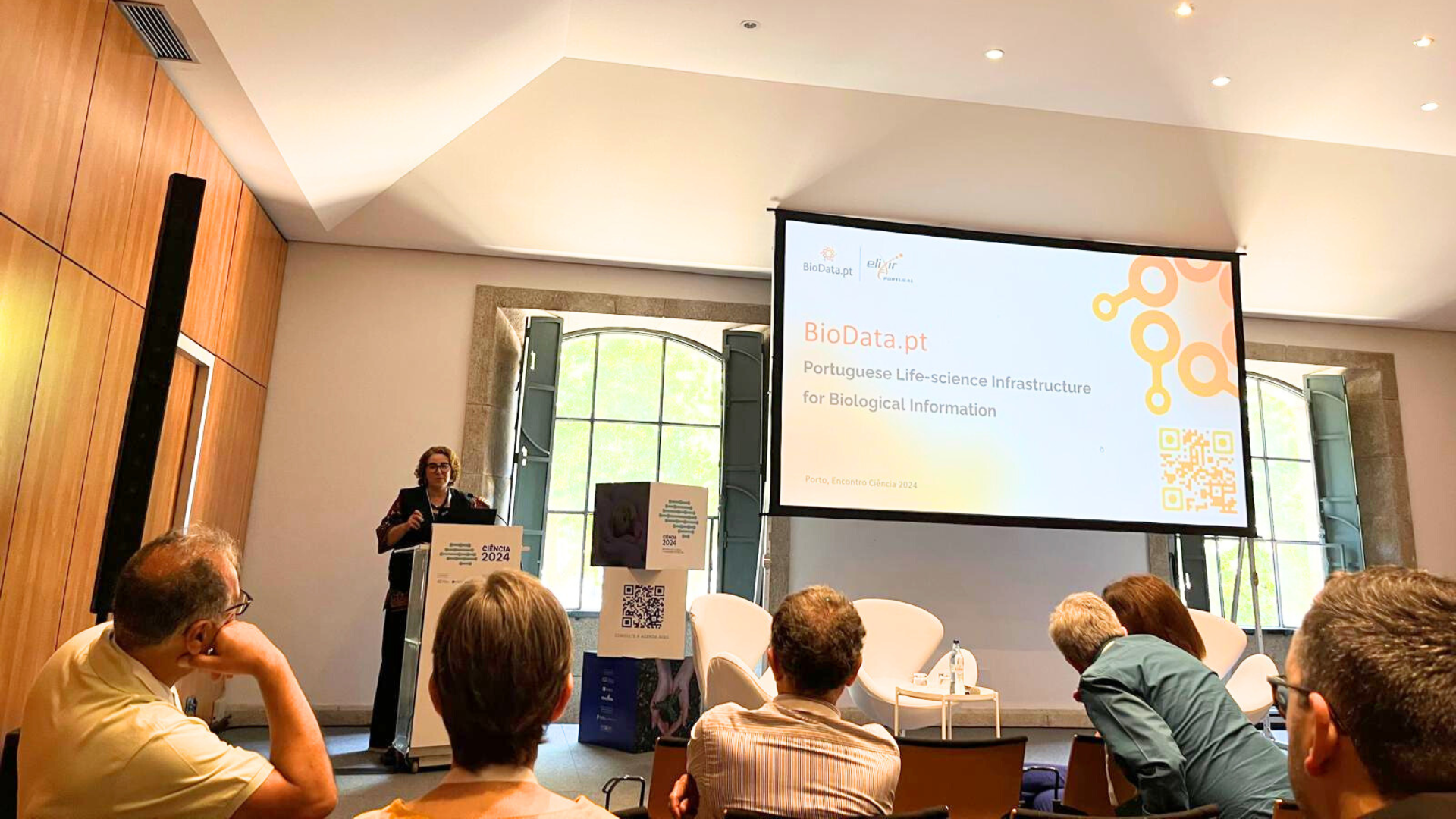 A presenter speaks at the Encontro Ciência 2024 in Porto, discussing BioData.pt, a Portuguese life-science infrastructure for biological information. Audience members listen attentively.